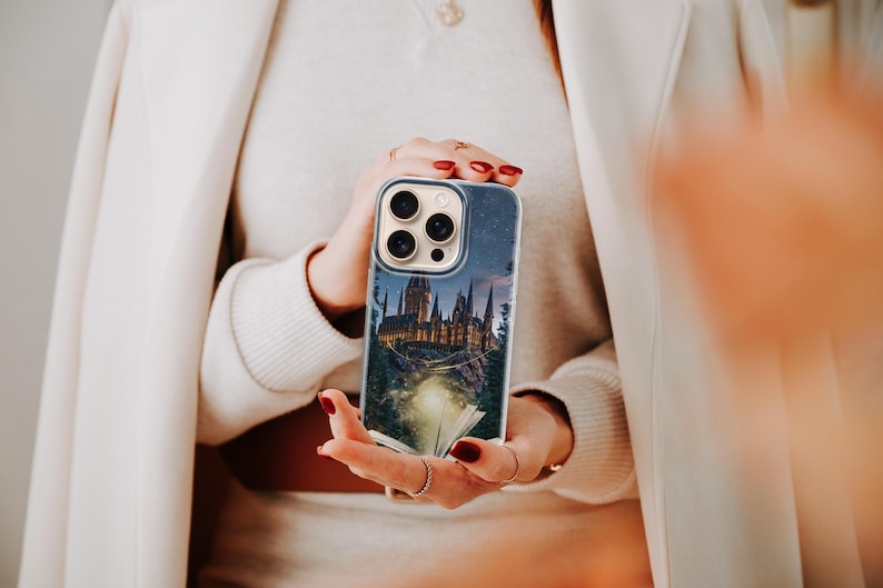 May include: A clear phone case featuring a nighttime castle scene with a starry sky. The case is held in front of a person wearing a cream-colored sweater and a white coat. The phone case has a camera and flash cutout.