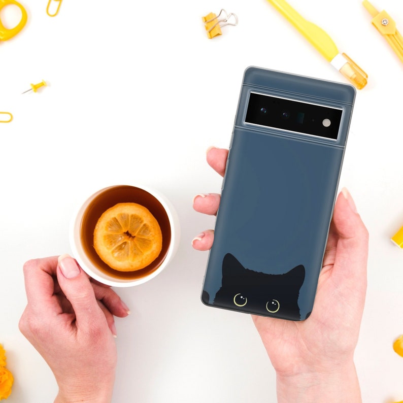 May include: A dark blue phone case with a black cat silhouette and yellow eyes. The cat is peeking out from behind the case.