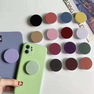 May include: A collection of colorful phone grips in various shades, including black, pink, blue, and yellow. Two smartphones, one lavender and one green, each have a matching grip attached. The grips are round and provide a secure hold.