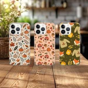 Op de afbeelding: Drie telefoonhoesjes met herfstthema's. Eén hoesje heeft pompoenen en bladeren, een ander heeft paddenstoelen en bessen, en de derde toont vossen, mokken en truien. De hoesjes liggen op een houten oppervlak.