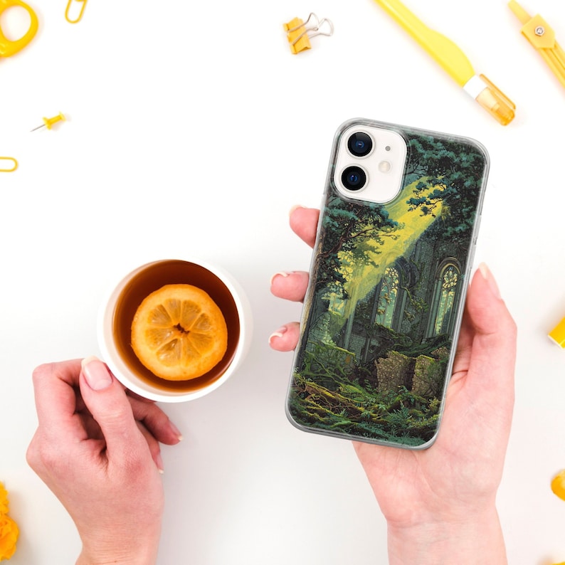 May include: A clear phone case with a green and yellow landscape design featuring a stone building with windows. The case is held in a hand.