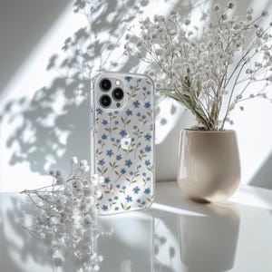 Op de afbeelding: Transparante telefoonhoes met een bloemenpatroon van kleine blauwe bloemen en groene stelen. De hoes wordt getoond met een witte bloemarrangement in een beige vaas. De hoes ligt op een wit oppervlak met schaduwen.