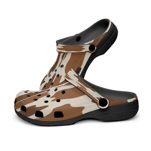 May include: A pair of clogs with a brown and cream cow print pattern. The clogs have a black sole and strap, and feature multiple ventilation holes. The shoes are designed for comfort and casual wear.