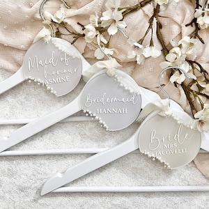 Könnte beinhalten: Weiße Holzkleiderbügel mit runden, mattierten Namensschildern aus Acryl. Jedes Schild ist mit Kunstperlen verziert und trägt eleganten Schriftzug: „Maid of Honour Jasmine“, „Bridesmaid Hannah“ und „Bride Mrs. Jacovelli“. Bänder und florale Akzente runden das Design ab.