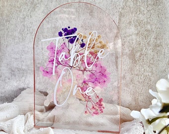 May include: Clear acrylic table number holder with a rose gold top. The holder features dried flowers in shades of pink, purple, and white. The text "Table One" is written in white on the front of the holder.