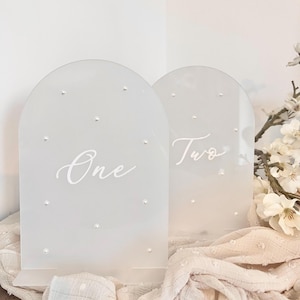 May include: Two frosted acrylic table numbers, arched at the top, with the numbers "One" and "Two" in elegant script. Each number is adorned with small, sparkling embellishments. The table numbers are displayed on a cream-colored surface.