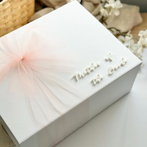K&ouml;nnte beinhalten: Eine wei&szlig;e Geschenkbox mit einer hellrosa T&uuml;llschleife. Auf der Oberseite der Box steht in wei&szlig;er Schrift "Mother of the Bride". Die Box steht auf einer wei&szlig;en Oberfl&auml;che.