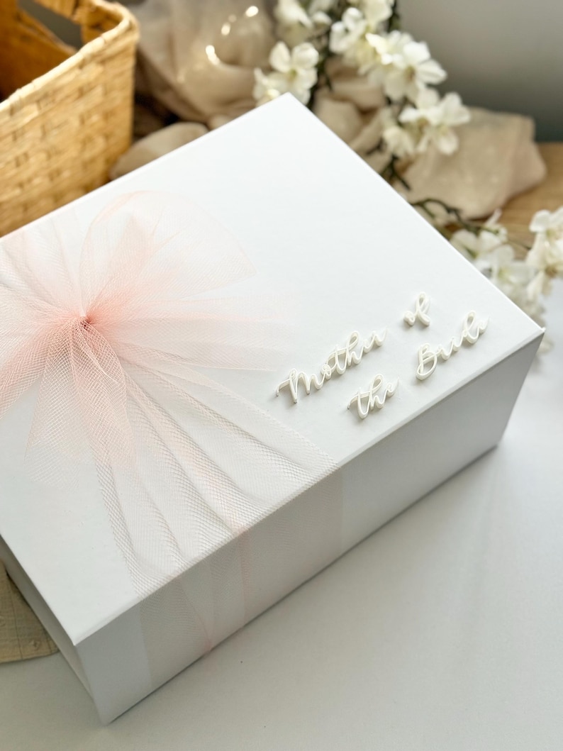 K&ouml;nnte beinhalten: Eine wei&szlig;e Geschenkbox mit einer hellrosa T&uuml;llschleife. Auf der Oberseite der Box steht in wei&szlig;er Schrift "Mother of the Bride". Die Box steht auf einer wei&szlig;en Oberfl&auml;che.