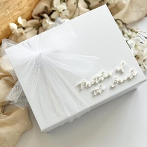 K&ouml;nnte beinhalten: Eine wei&szlig;e Geschenkbox mit dem Schriftzug "Mother of the Bride" in Schreibschrift. Die Box ist mit einer durchsichtigen wei&szlig;en Schleife verziert. Der Hintergrund umfasst wei&szlig;e Blumen und einen Weidenkorb, was auf ein Hochzeitsthema hindeutet.