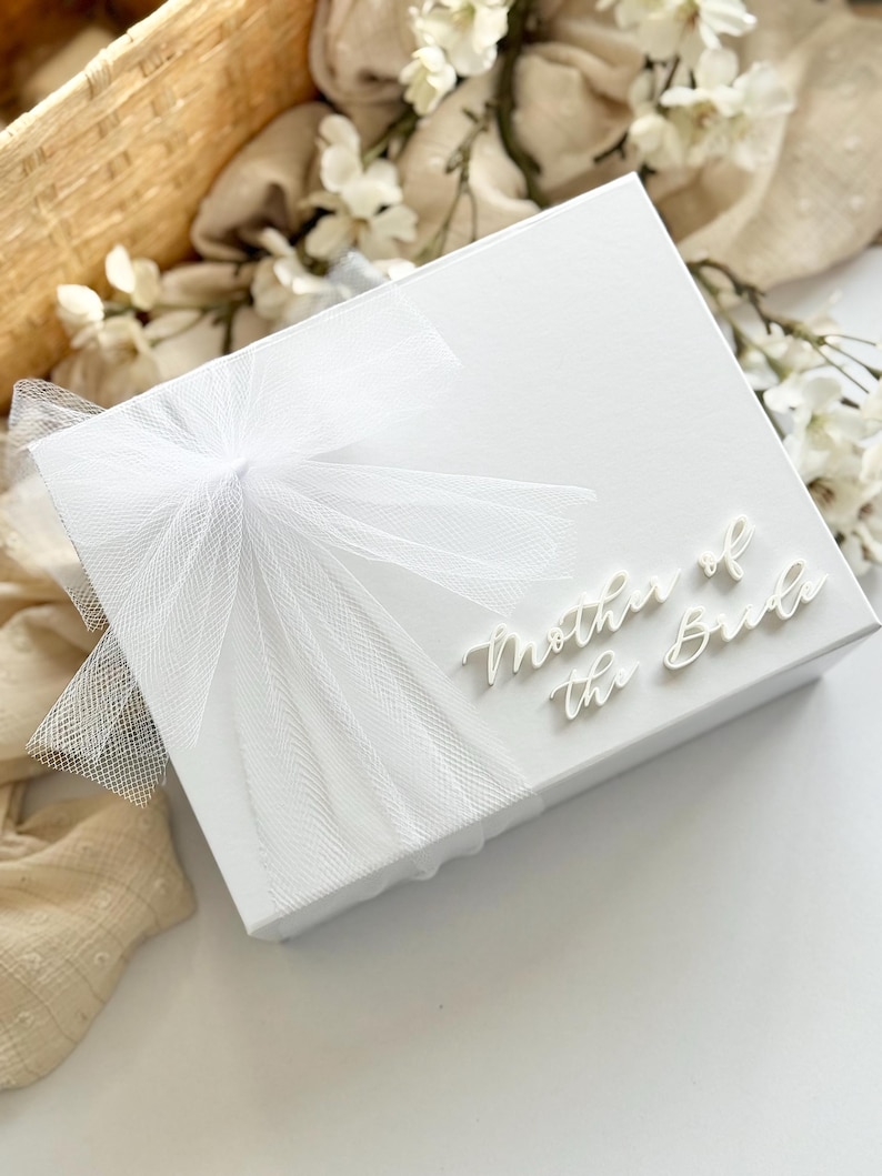 K&ouml;nnte beinhalten: Eine wei&szlig;e Geschenkbox mit dem Schriftzug "Mother of the Bride" in Schreibschrift. Die Box ist mit einer durchsichtigen wei&szlig;en Schleife verziert. Der Hintergrund umfasst wei&szlig;e Blumen und einen Weidenkorb, was auf ein Hochzeitsthema hindeutet.