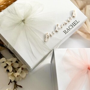 May include: White gift boxes with tulle bows. One box has "Bridesmaid RACHEL" in script. Another box has a pink tulle bow. The boxes are on a white surface with white flowers.