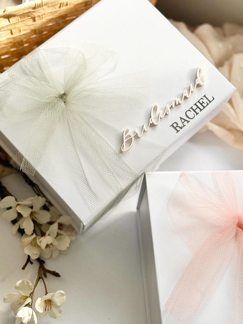 May include: White gift boxes with tulle bows. One box has "Bridesmaid RACHEL" in script. Another box has a pink tulle bow. The boxes are on a white surface with white flowers.