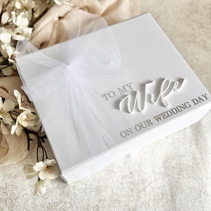 May include: A white gift box with the words "TO MY WIFE ON OUR WEDDING DAY" in silver lettering. The box is adorned with a delicate white tulle bow. White flowers and fabric are in the background.