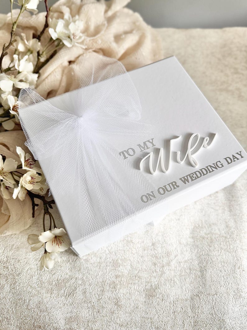 May include: A white gift box with the words "TO MY WIFE ON OUR WEDDING DAY" in silver lettering. The box is adorned with a delicate white tulle bow. White flowers and fabric are in the background.
