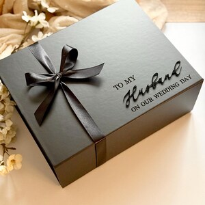 Puede incluir: Una caja de regalo gris oscuro con una cinta de sat&eacute;n negra atada con un lazo. La caja est&aacute; inscrita con las palabras "TO MY Husband ON OUR WEDDING DAY" en una elegante escritura. Un regalo perfecto para una boda.