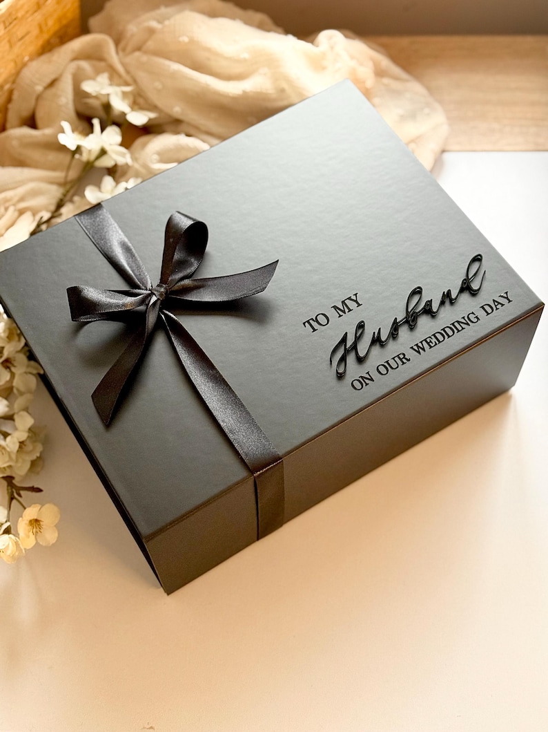 Puede incluir: Una caja de regalo gris oscuro con una cinta de sat&eacute;n negra atada con un lazo. La caja est&aacute; inscrita con las palabras "TO MY Husband ON OUR WEDDING DAY" en una elegante escritura. Un regalo perfecto para una boda.