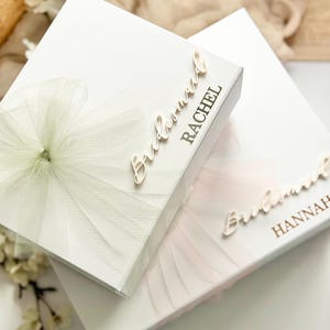 May include: Two white gift boxes, one with a light green tulle bow and the name "Rachel" in gold script, and the other with a pink tulle bow and the name "Hannah". The boxes are likely for a bridal party.