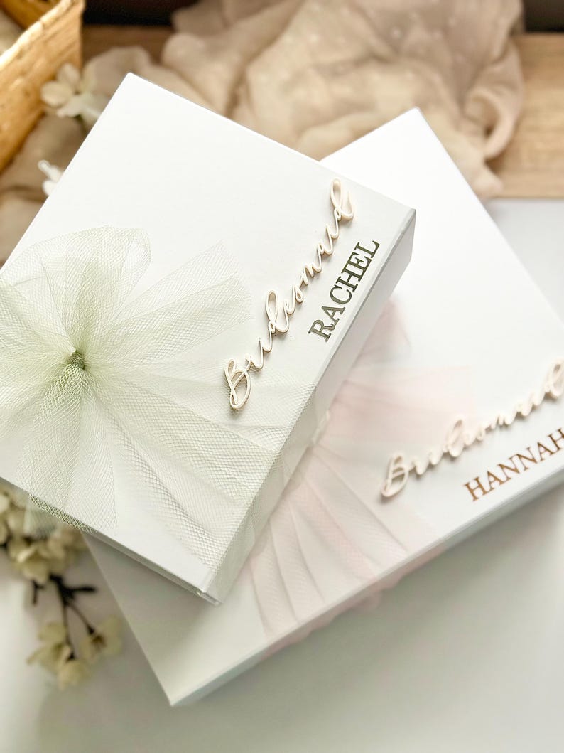 May include: Two white gift boxes, one with a light green tulle bow and the name "Rachel" in gold script, and the other with a pink tulle bow and the name "Hannah". The boxes are likely for a bridal party.