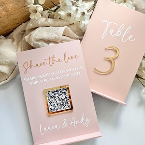 May include: Two pink table signs with white and gold lettering. One sign reads "Share the love" and includes a QR code. The other sign reads "Table 3" with a gold number. Floral arrangement in the background.