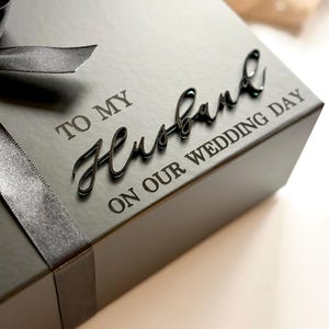 Puede incluir: Una caja de regalo negra con una cinta de raso. La caja tiene las palabras "TO MY Husband ON OUR WEDDING DAY" en una fuente de escritura. La caja est&aacute; sobre una superficie blanca.