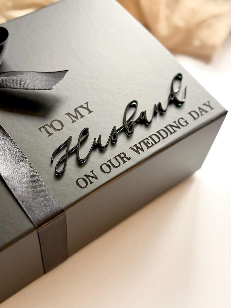 Puede incluir: Una caja de regalo negra con una cinta de raso. La caja tiene las palabras "TO MY Husband ON OUR WEDDING DAY" en una fuente de escritura. La caja est&aacute; sobre una superficie blanca.