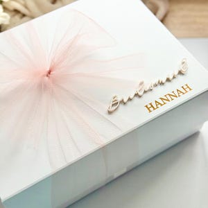 May include: A white gift box with a large, light pink tulle bow. The box is decorated with the word "Bridesmaid" in cursive and the name "HANNAH" in gold lettering. The box is likely for a wedding.