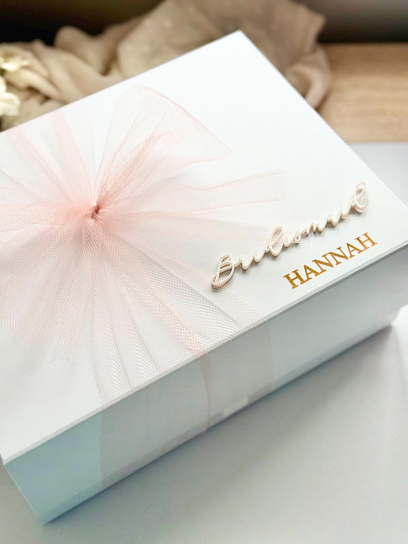May include: A white gift box with a large, light pink tulle bow. The box is decorated with the word "Bridesmaid" in cursive and the name "HANNAH" in gold lettering. The box is likely for a wedding.