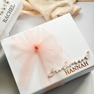 May include: White gift box with a sheer peach bow and the word "Bridesmaid" and the name "Hannah" in gold lettering. Another box with the name "Rachel" is partially visible. The background includes a cream-colored fabric.