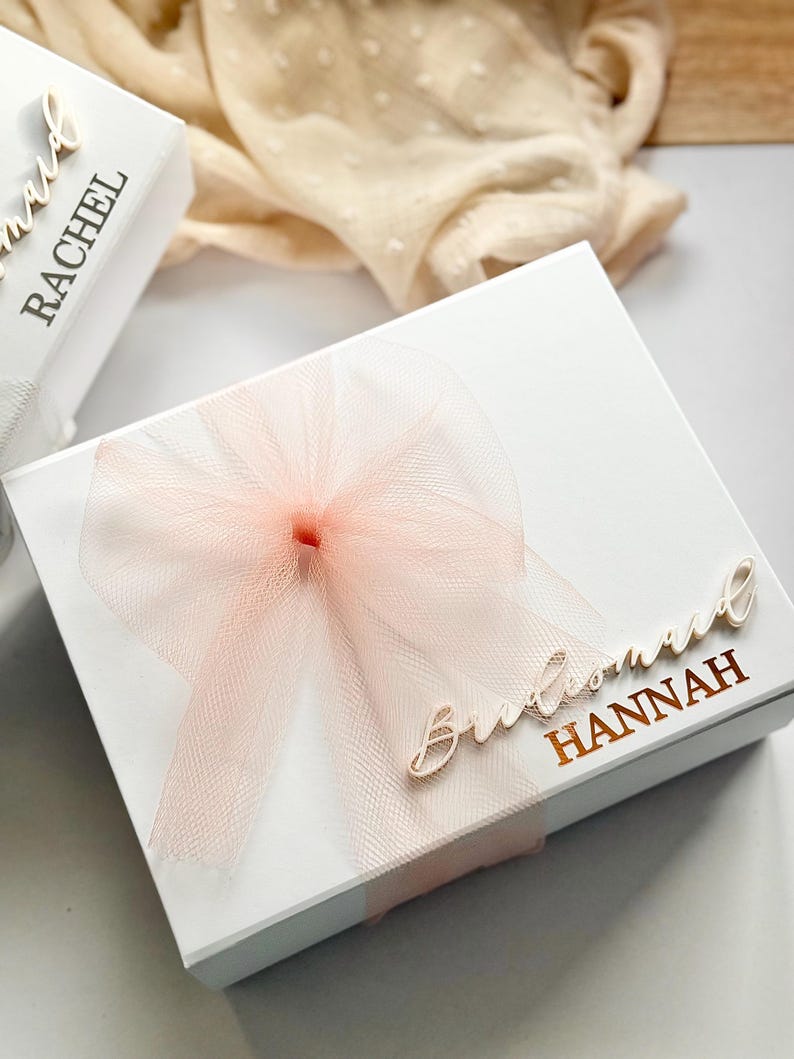 May include: White gift box with a sheer peach bow and the word "Bridesmaid" and the name "Hannah" in gold lettering. Another box with the name "Rachel" is partially visible. The background includes a cream-colored fabric.
