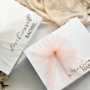 May include: Two white gift boxes with tulle bows. One box has a white bow, the other a pink bow. The boxes are labeled "Bridesmaid" and a name. The boxes are likely for a wedding or bridal party.