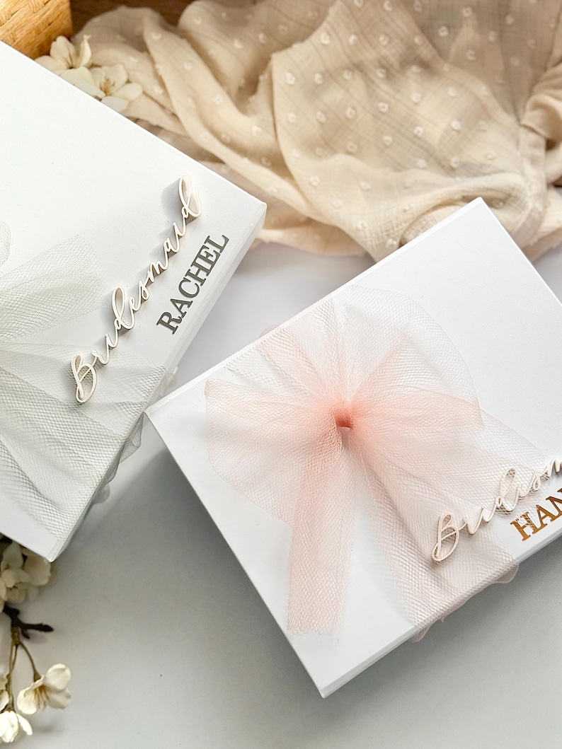 May include: Two white gift boxes with tulle bows. One box has a white bow, the other a pink bow. The boxes are labeled "Bridesmaid" and a name. The boxes are likely for a wedding or bridal party.