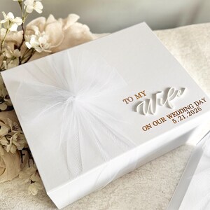 May include: A white gift box with a sheer white ribbon and bow. The box has the words "TO MY WIFE ON OUR WEDDING DAY 5.21.2026" in gold lettering. The box is on a white surface.