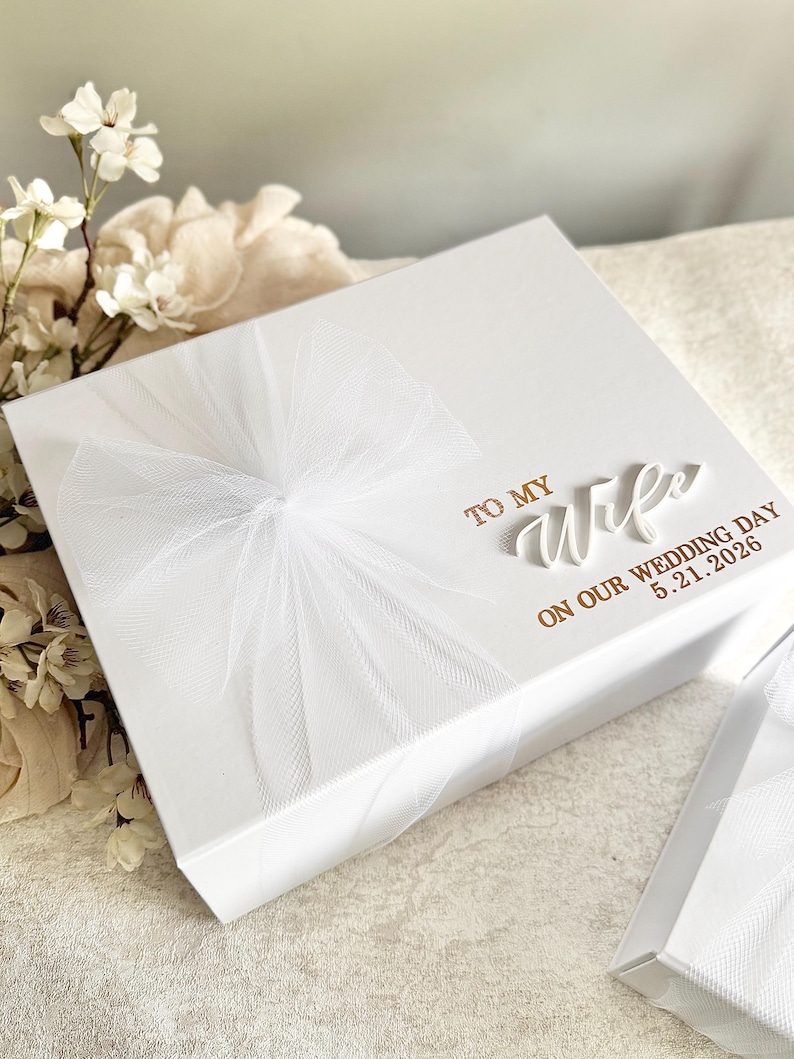 May include: A white gift box with a sheer white ribbon and bow. The box has the words "TO MY WIFE ON OUR WEDDING DAY 5.21.2026" in gold lettering. The box is on a white surface.