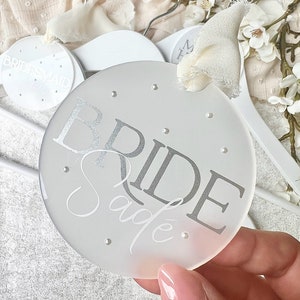 May include: White frosted round hanger with the word "BRIDE" in silver lettering and the name "Sade" written in cursive below. The hanger has small silver dots around the edge.
