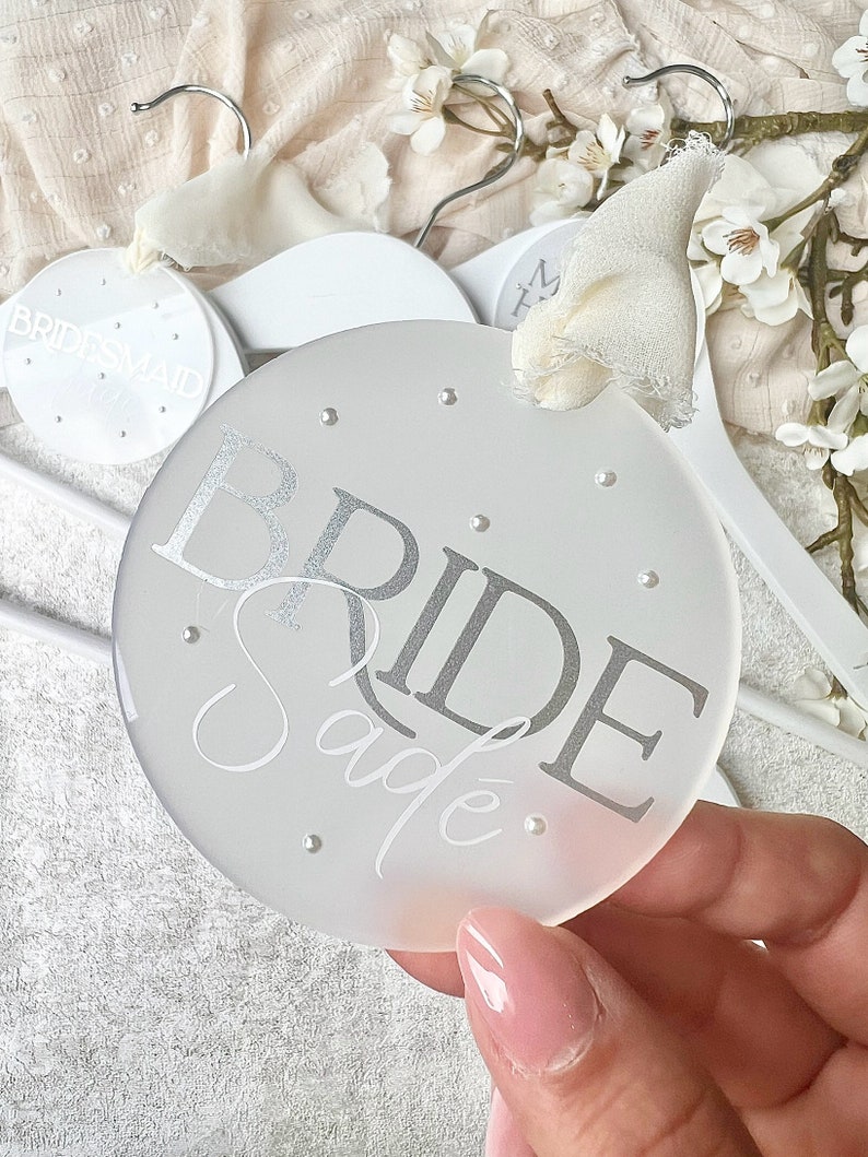 May include: White frosted round hanger with the word "BRIDE" in silver lettering and the name "Sade" written in cursive below. The hanger has small silver dots around the edge.