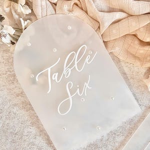Könnte beinhalten: Weißes Acryl-Tischnummernschild mit dem Text "Table Six" in Schreibschrift. Das Schild ist mit kleinen weißen Perlen verziert.