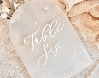May include: White acrylic table number sign with the text "Table Six" in a script font. The sign is decorated with small white pearl-like embellishments.