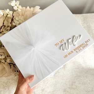 May include: A white gift box with a tulle bow and gold lettering that reads "TO MY Wife ON OUR WEDDING DAY 5.21.2026". The box is held by a person.