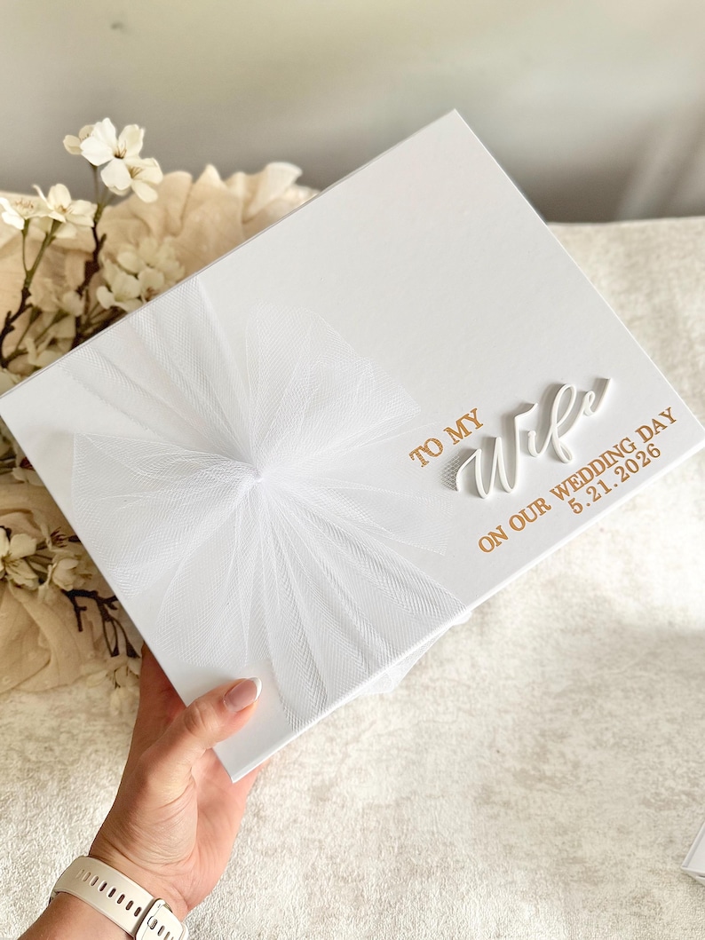 May include: A white gift box with a tulle bow and gold lettering that reads "TO MY Wife ON OUR WEDDING DAY 5.21.2026". The box is held by a person.