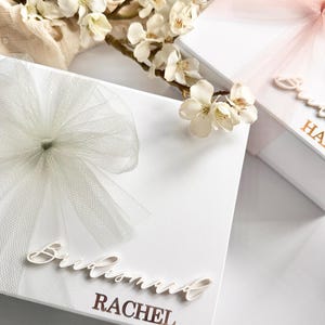 May include: White gift boxes with tulle bows and floral accents. One box has "Bridesmaid RACHEL" in cursive script. Another box has "Sweet HA" in gold lettering. The boxes are decorated with white and pink tulle bows.