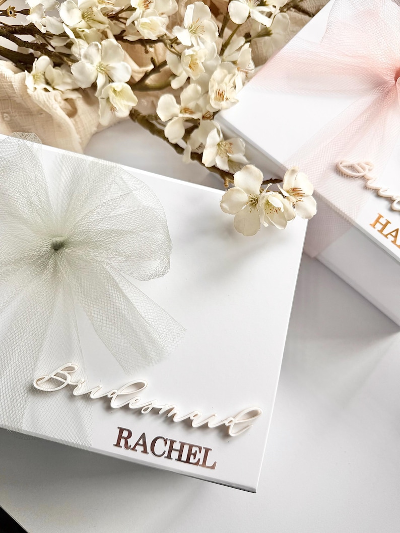 May include: White gift boxes with tulle bows and floral accents. One box has "Bridesmaid RACHEL" in cursive script. Another box has "Sweet HA" in gold lettering. The boxes are decorated with white and pink tulle bows.