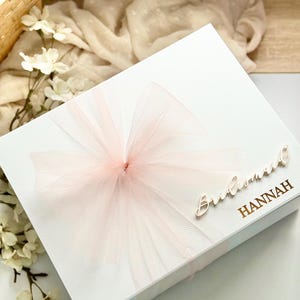 May include: A white gift box with a large, light pink tulle bow. The box is adorned with the word "Bridesmaid" and the name "Hannah" in a gold script. The box is decorated with white flowers.