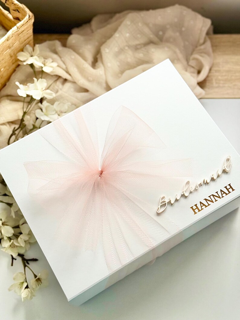 May include: A white gift box with a large, light pink tulle bow. The box is adorned with the word "Bridesmaid" and the name "Hannah" in a gold script. The box is decorated with white flowers.