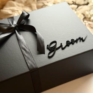 May include: A black gift box tied with a black satin ribbon and bow. The word "Groom" is written in cursive on the box. Ideal for a wedding or special occasion.