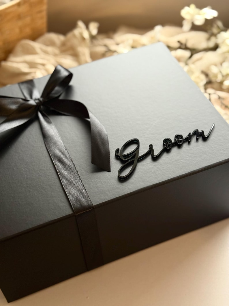 May include: A black gift box tied with a black satin ribbon and bow. The word "Groom" is written in cursive on the box. Ideal for a wedding or special occasion.