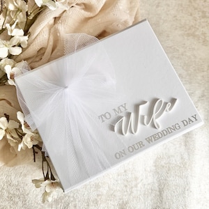 May include: A white wedding keepsake box with the words "TO MY Wife ON OUR WEDDING DAY" in silver lettering. The box is adorned with a white tulle bow. White flowers and a cream-colored fabric are in the background.