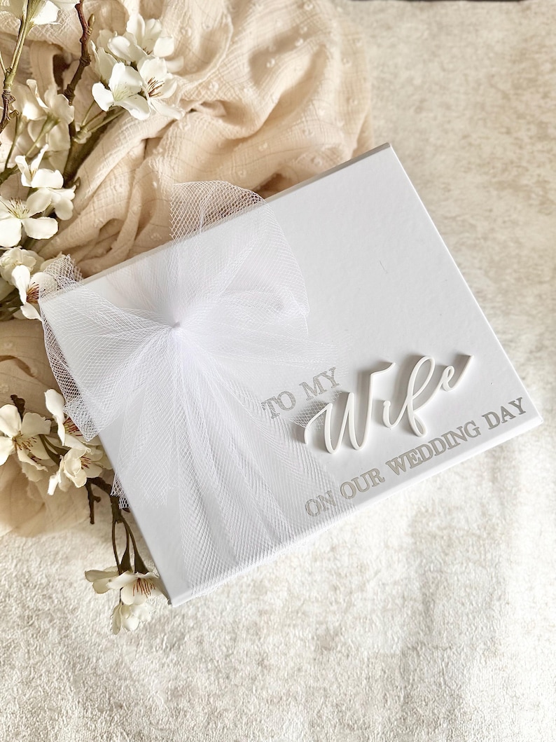 May include: A white wedding keepsake box with the words "TO MY Wife ON OUR WEDDING DAY" in silver lettering. The box is adorned with a white tulle bow. White flowers and a cream-colored fabric are in the background.