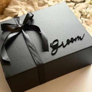 May include: A black gift box tied with a black satin ribbon and bow. The word "Groom" is written in a cursive font on the top of the box. The box is on a light-coloured surface.