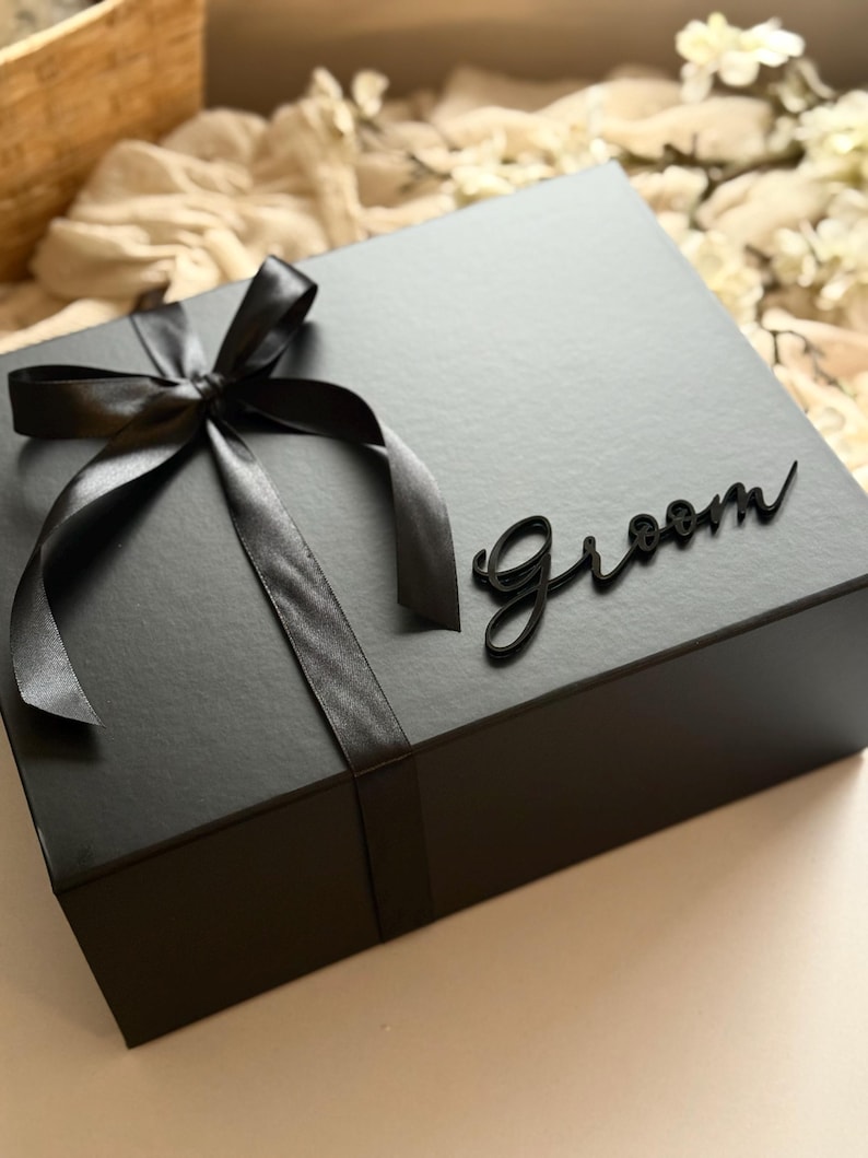 May include: A black gift box tied with a black satin ribbon and bow. The word "Groom" is written in a cursive font on the top of the box. The box is on a light-coloured surface.