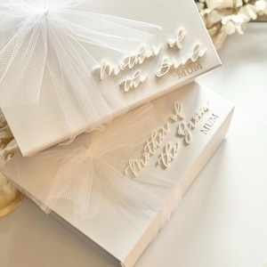 K&ouml;nnte beinhalten: Zwei wei&szlig;e Geschenkboxen mit den Schriftz&uuml;gen "Mother of the Bride" und "Mother of the Groom" in Schreibschrift. Jede Box ist mit einer wei&szlig;en T&uuml;llschleife verziert. Das Wort "MUM" ist darunter gedruckt.